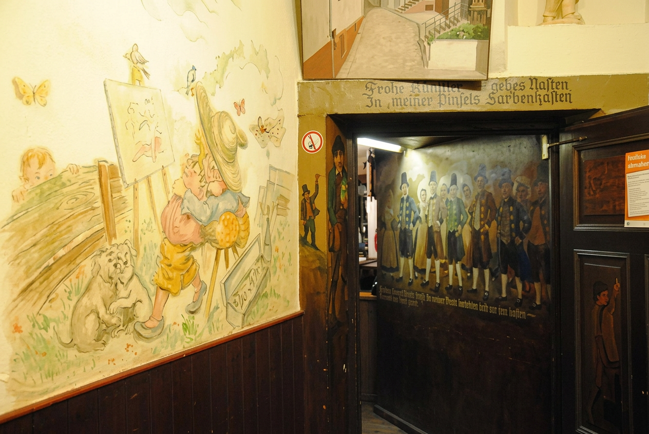 OPTIMIERT_Ein Blick in den Eingangsbereich des Malkastens mit den Malereien von Joachim Liebich und der von ihm geschaffenen Figur des MEISTER PINSEL, vorne links. Auf malerkolonie-schwalenbach.de-gadthoefe-malkasten 17.10.20